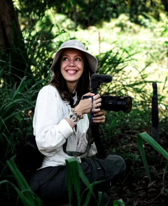 A woman in a white shirt and hat is sitting in the woods.